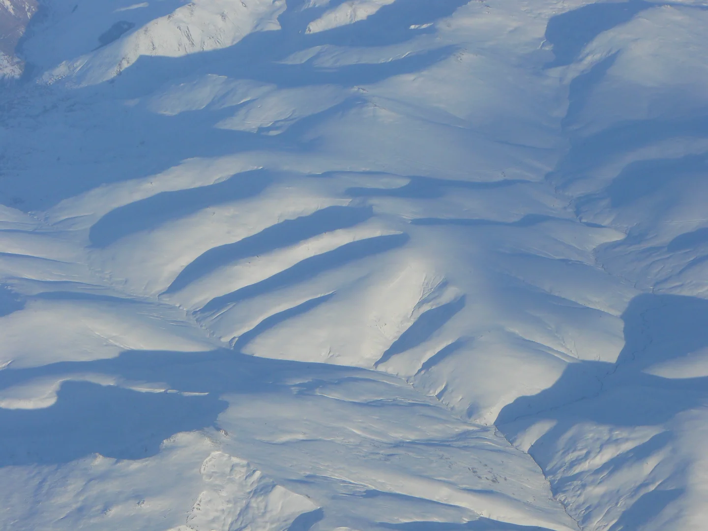Snow-covered landscape with ridged terrain and distant mountain peaks under clear blue sky.