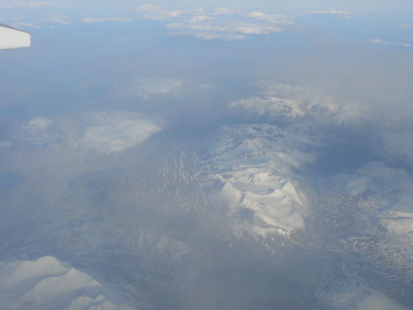Snowy mountains stretch beneath a hazy sky, with an airplane wing visible in the corner.