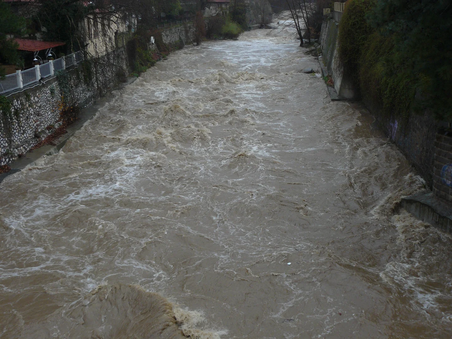 Muddy river flows swiftly between stone embankments in an urban setting.