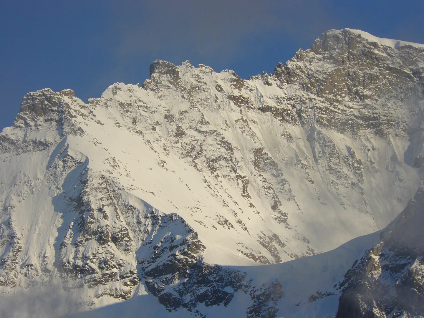 Steep snow-covered mountain face with grey rock outcrops, lit warmly by low sun, beneath a deep blue sky with a thin wisp of cloud.
