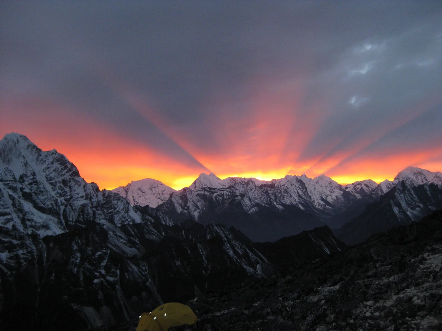 Rays of orange light fanning out across a grey sky behind a jagged line of snowy mountain peaks at sunset, with a yellow tent on a rocky slope in the foreground.
