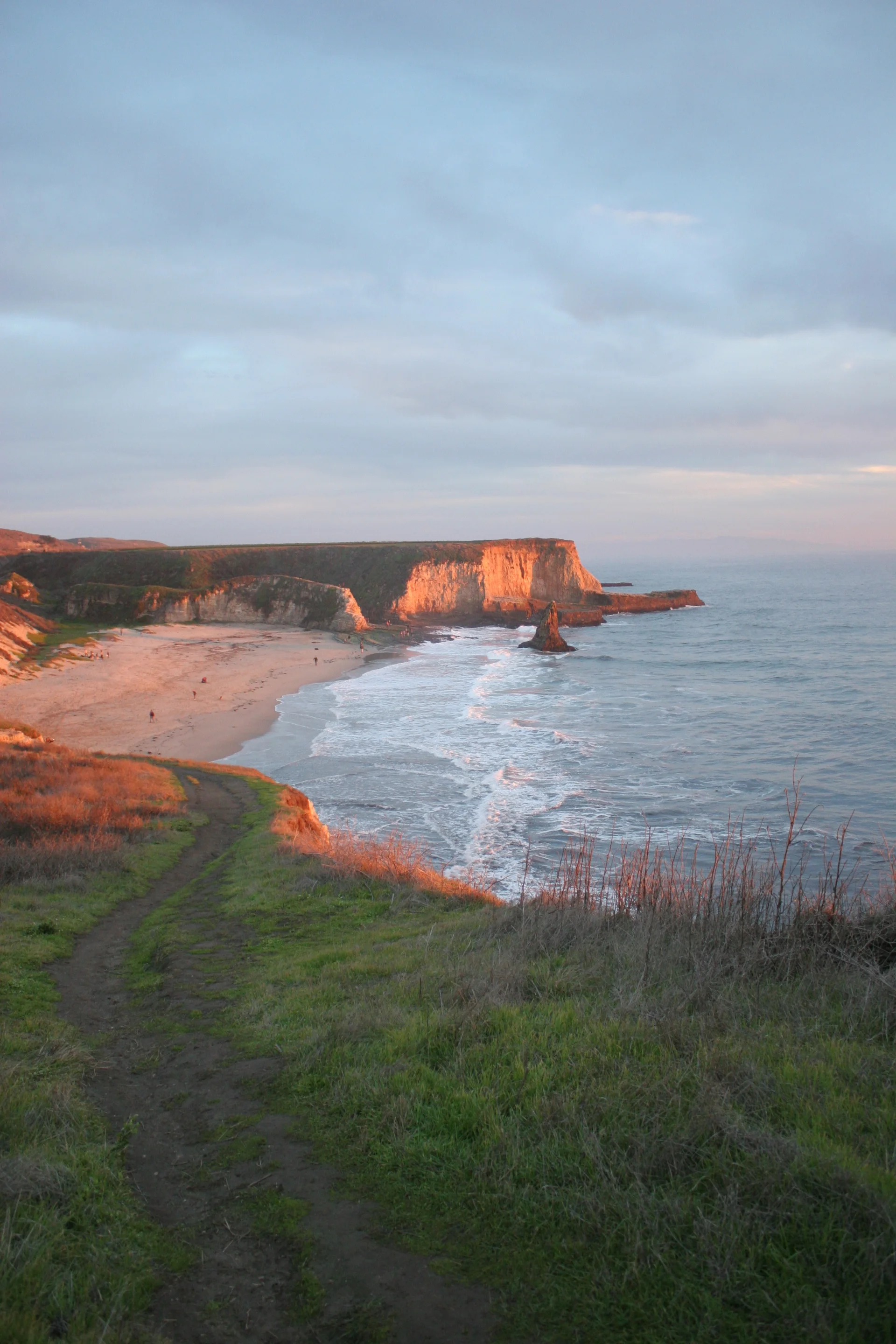 Cliffside beach with sandy shore, grassy path, and ocean under a partly cloudy sky.
