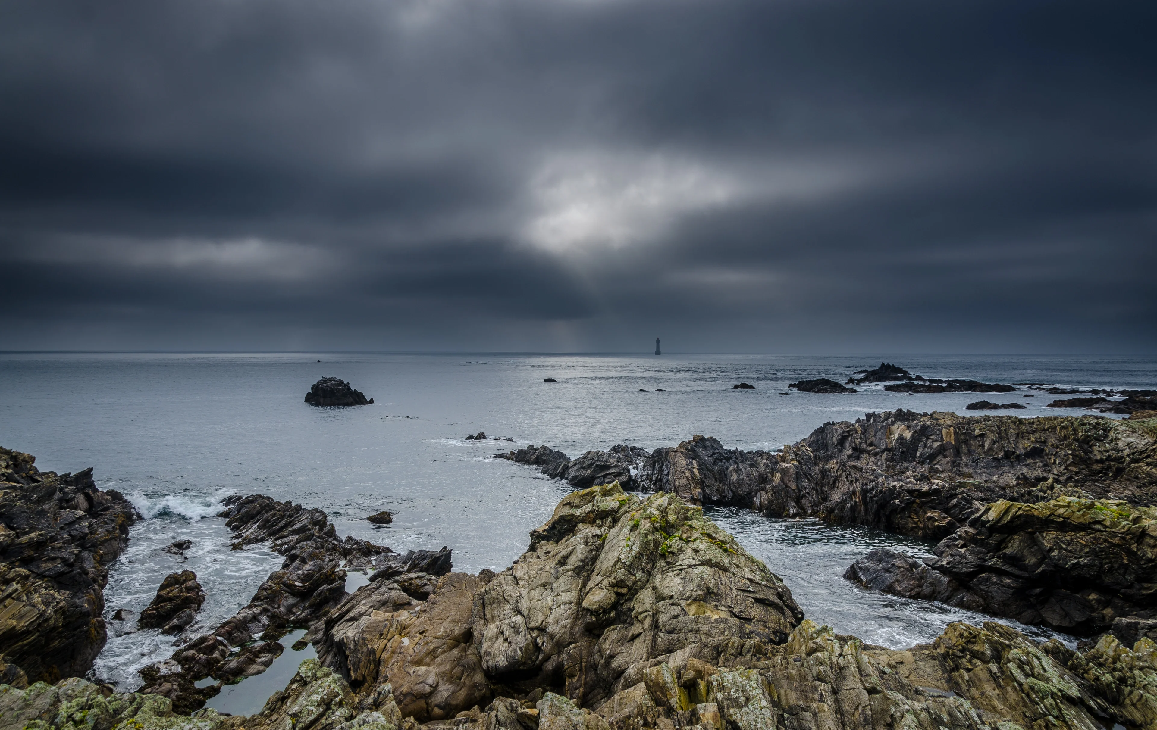 Rugged cliffs meet the sea under a stormy sky, with a lighthouse in the distance.