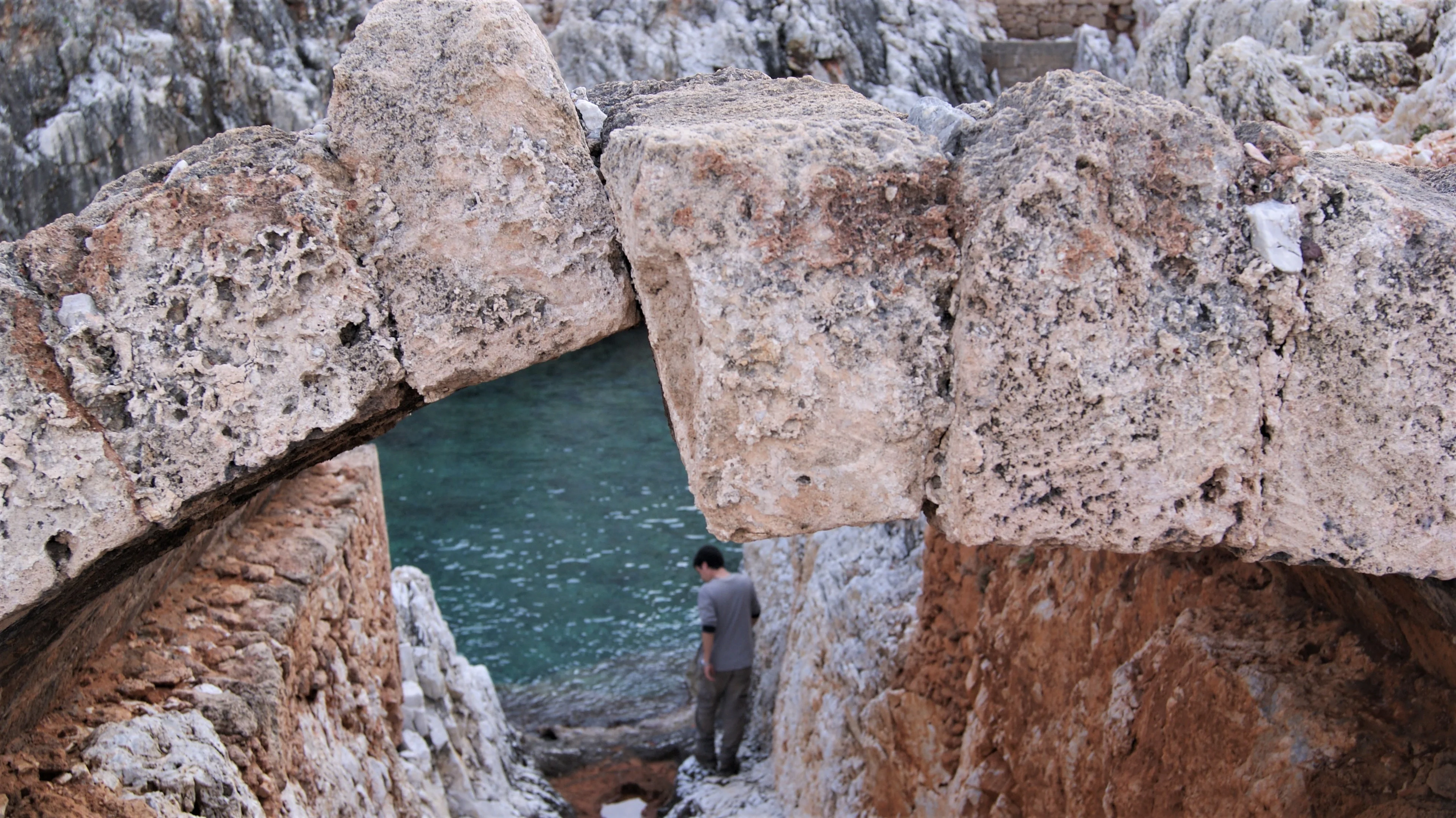 Person stands under ancient stone archway near calm sea.