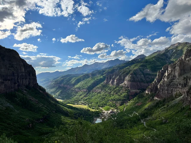 Deep green valley flanked by steep cliffs, with a small town and a pond on the valley floor and forested ridges and mountains beyond under a blue sky with white clouds.