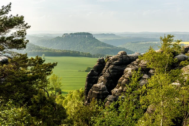 Weathered sandstone rock towers amid green trees, overlooking bright fields and a forested flat-topped hill crowned by fortress walls in the hazy distance.