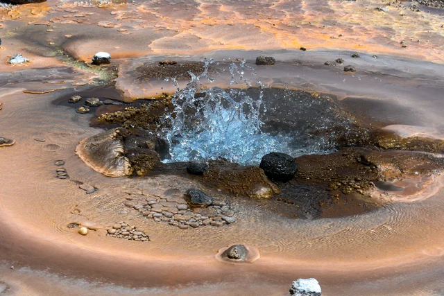 Small pool of clear water bubbling and splashing up from a hole, surrounded by orange, brown and dark crusted ground with scattered pebbles and stones.