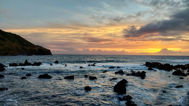 A serene sunset over the ocean, with rocks scattered in the shallow water and a cliff in the background.