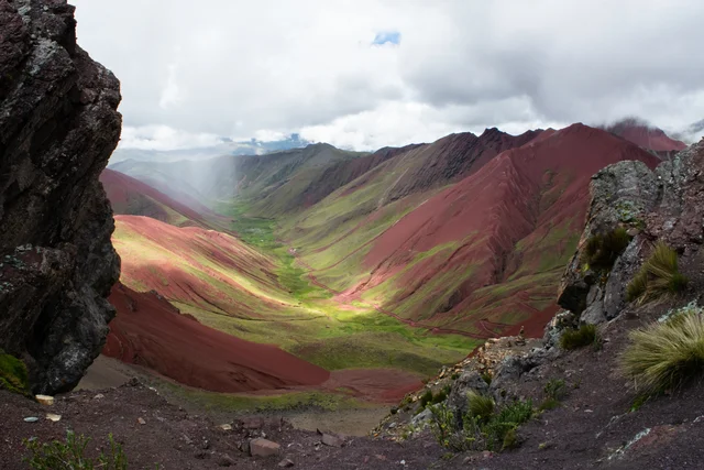 Valley between steep bright red slopes streaked with green grass, a winding red track along the floor, framed by dark rocks and tussock grass under grey cloud.