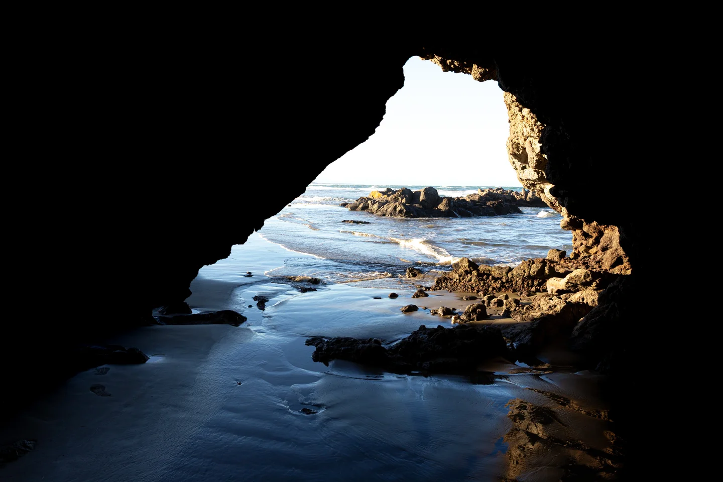 The photograph shows a rocky sea cave formed in volcanic rocks, opening onto a beach, with the dark cave interior contrasting strongly with the bright blue sea, breaking waves, wet sand, and sunlit rocks beyond.