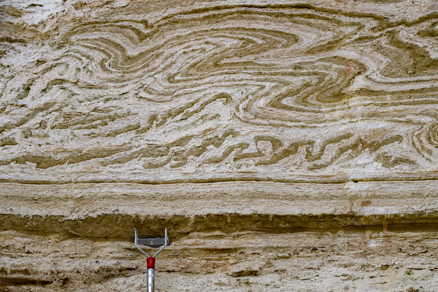 Layered sedimentary rock formation with swirling patterns and a red-handled tool at the bottom.