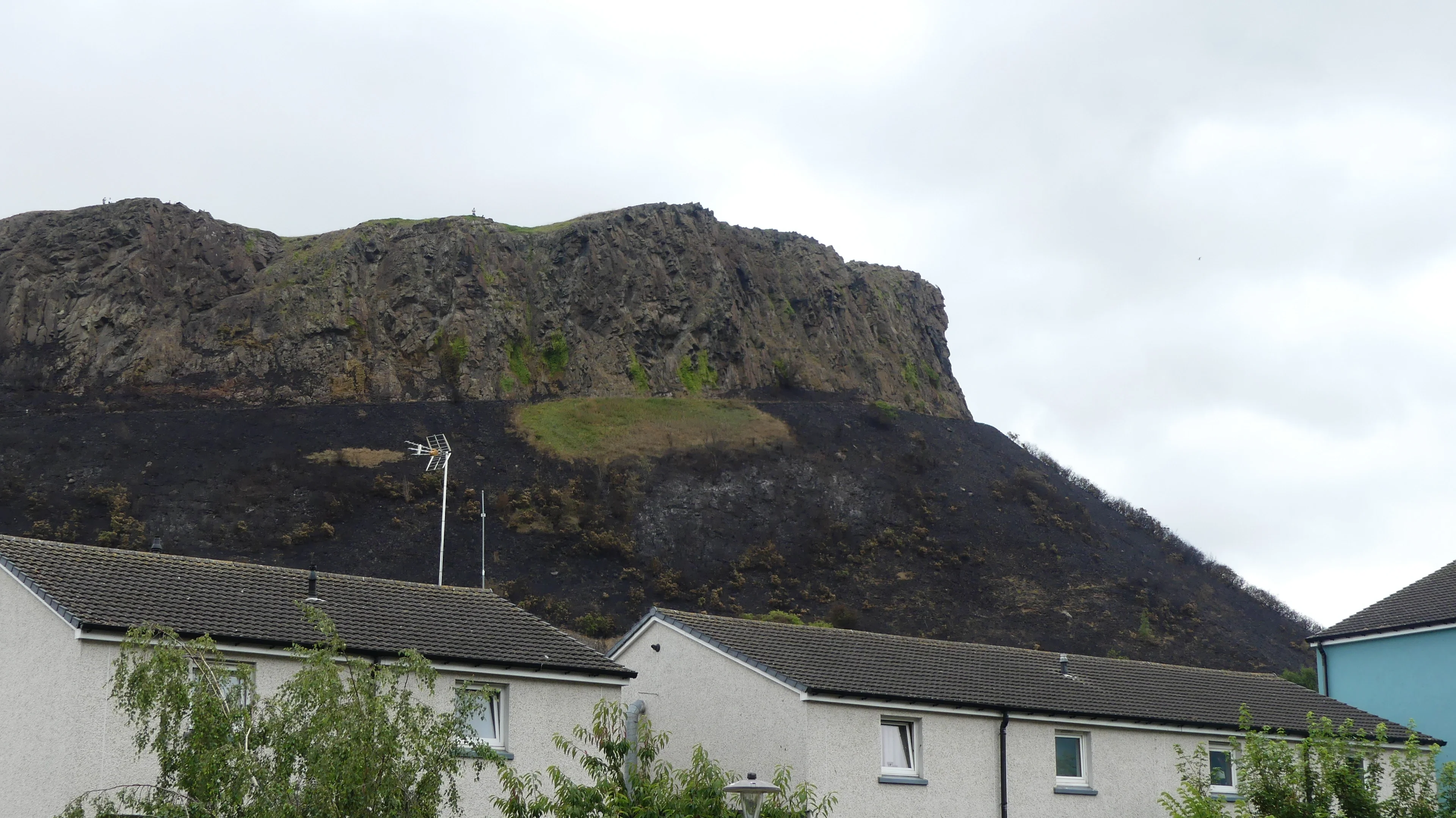A steep, rocky hillside dominates the scene, partially covered in dark, rough material. At its base, there are several residential houses with pitched roofs, and sparse green vegetation grows in front. The sky is overcast, creating a muted backdrop.