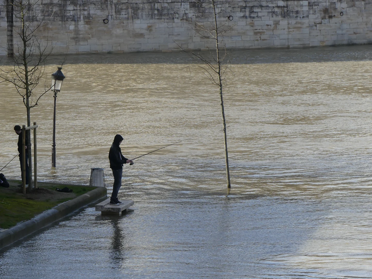 A person stands on a concrete platform near a flooded river, fishing with a rod. The scene includes bare trees, a lamppost, and a stone wall in the background. The water is calm, reflecting the grey sky.