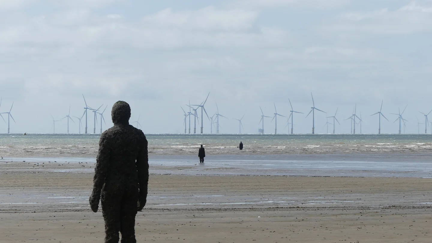 A weathered, textured figure stands silhouetted on a sandy shore, facing the ocean. In the distance, a line of wind turbines extends into the horizon under a cloudy sky. A person walks away towards the water, with another figure visible further out.