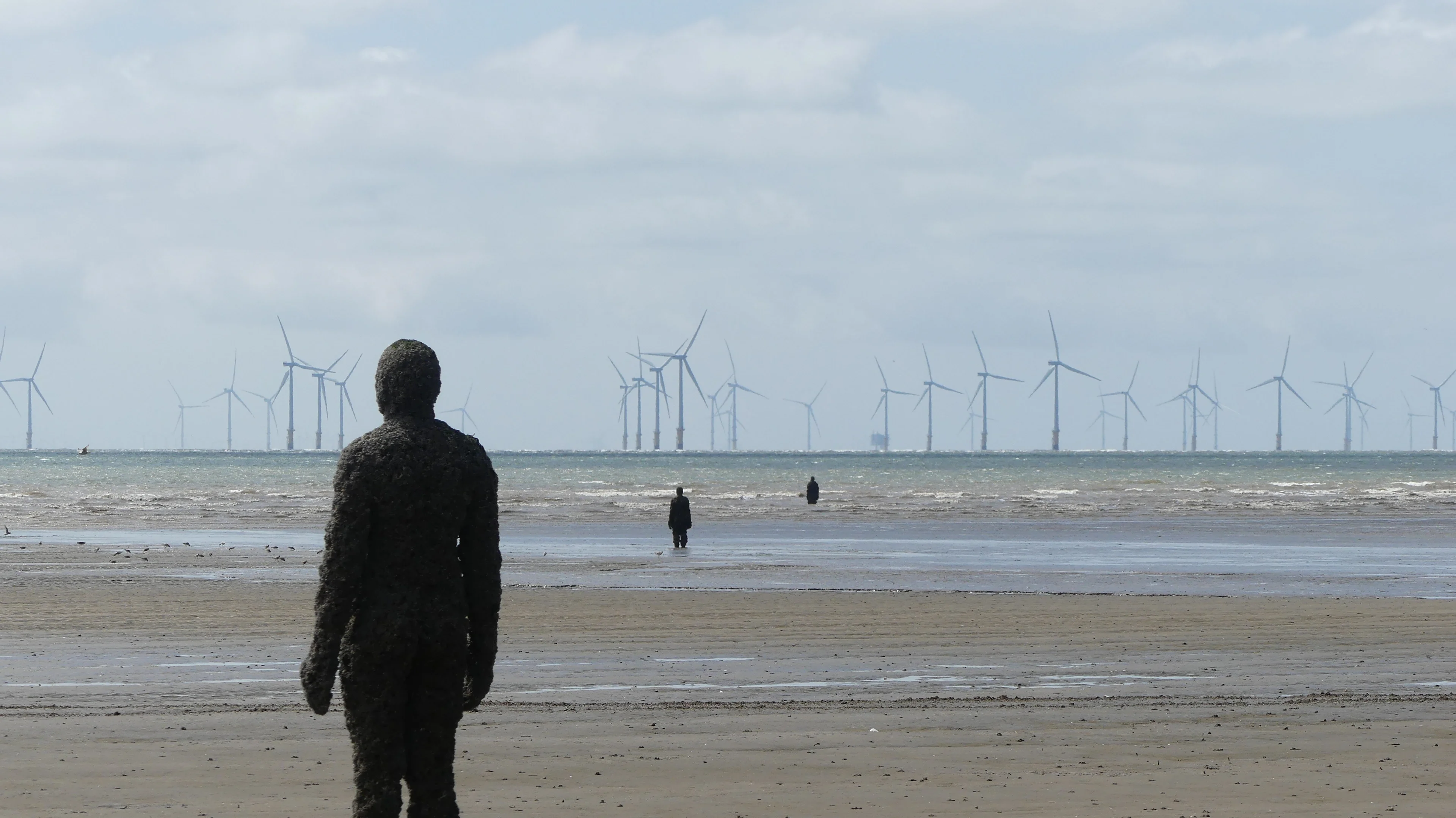 A weathered, textured figure stands silhouetted on a sandy shore, facing the ocean. In the distance, a line of wind turbines extends into the horizon under a cloudy sky. A person walks away towards the water, with another figure visible further out.